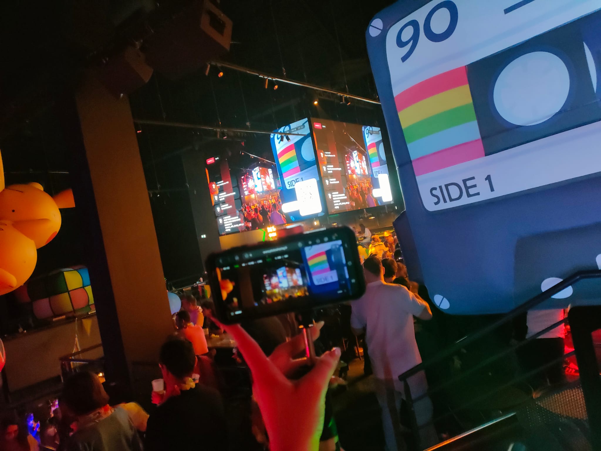Een smartphone filmt grote schermen met publieksfoto’s en QR-codes tijdens een kleurrijke retroavond.