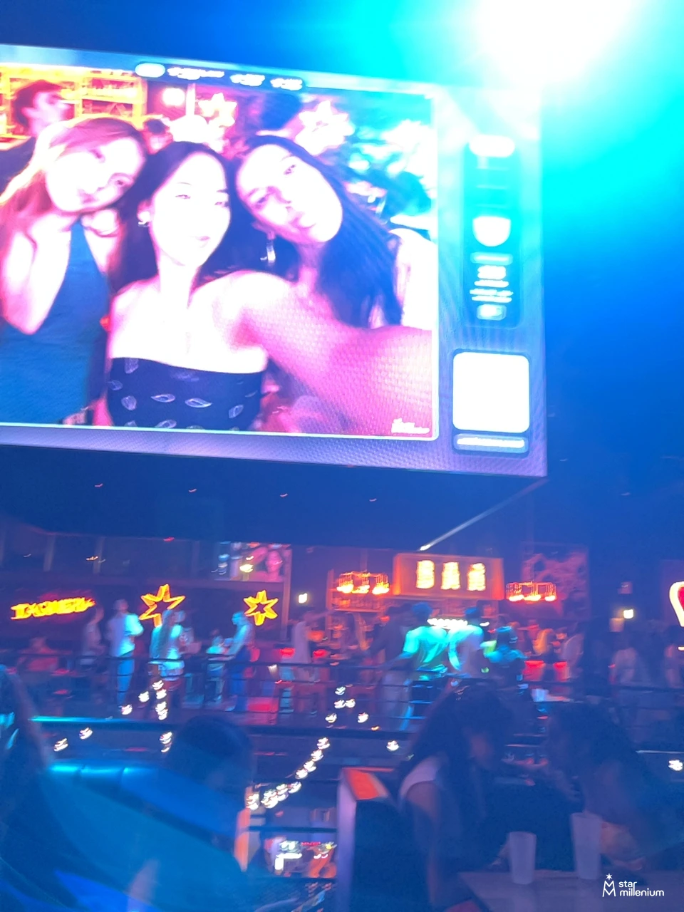 Grande schermo con il selfie di tre persone sopra una folla in un locale illuminato di blu e rosa