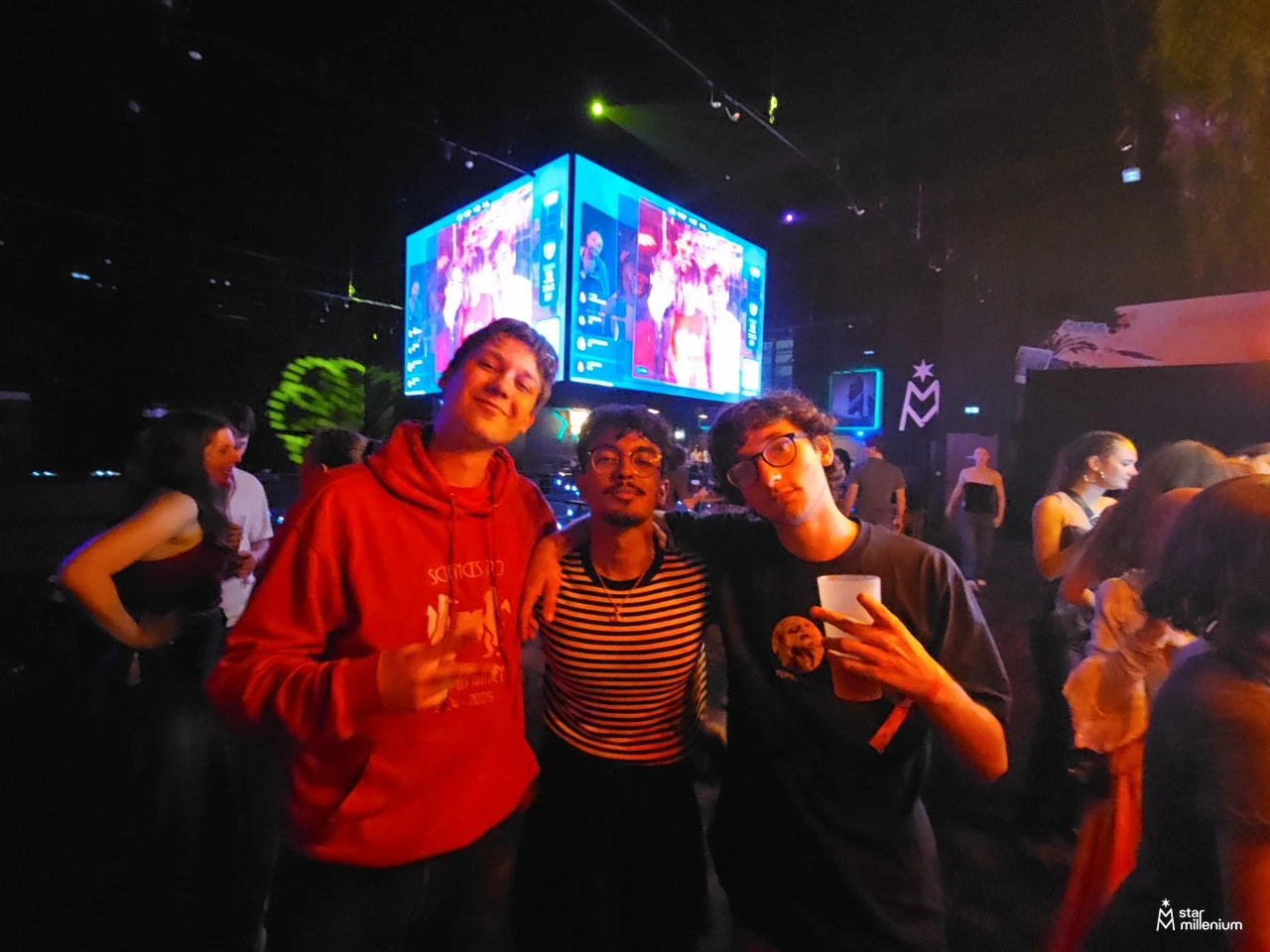 Three people posing with hand gestures and a white cup at an indoor party, with colorful suspended screens and stage lights behind them.