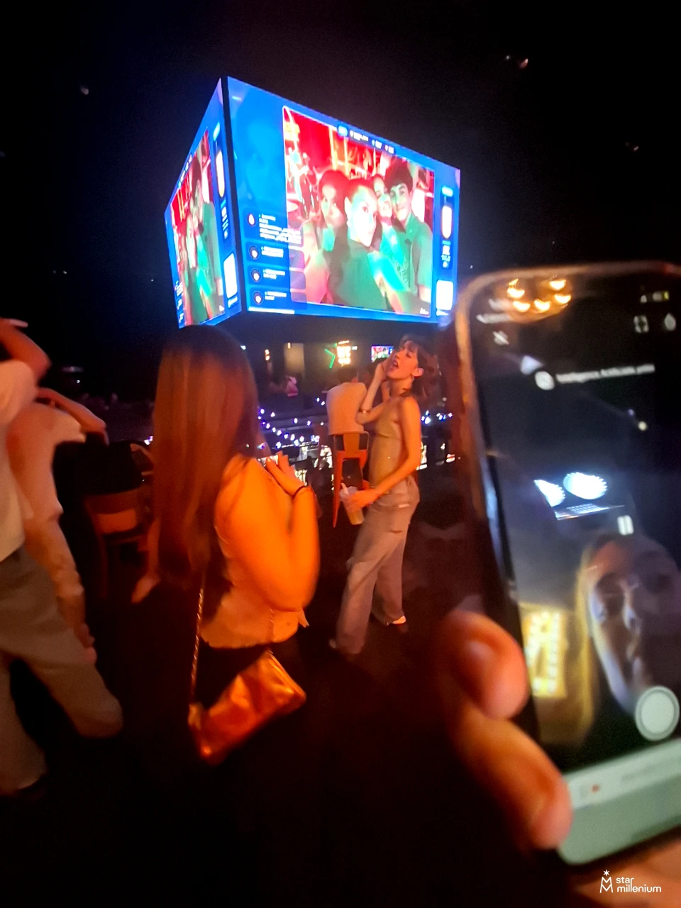 Partygoers filming a person who appears to be singing beneath a large illuminated screen in a dark venue