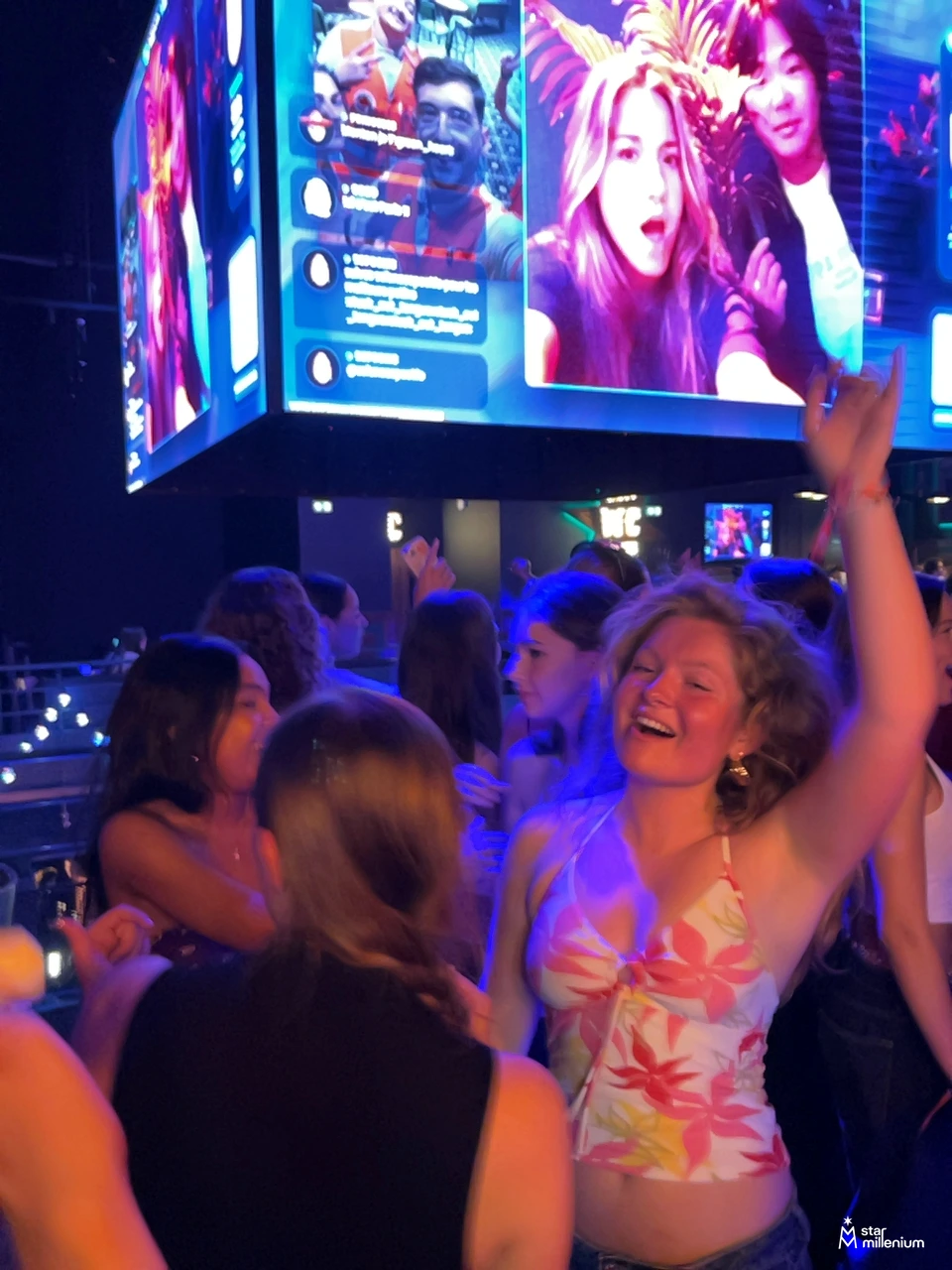 Smiling person dancing with one arm raised in a blue-and-pink-lit crowd beneath a large screen showing selfies and comments
