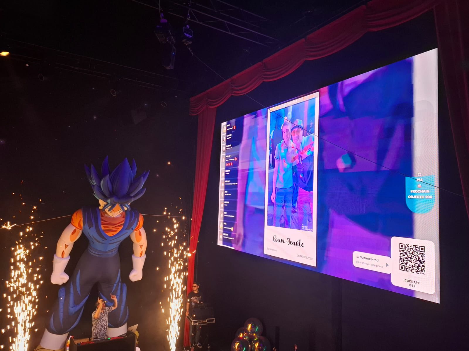 Indoor stage with spark fountains, a giant blue-haired character figure, two people and a large screen showing a photo and QR code