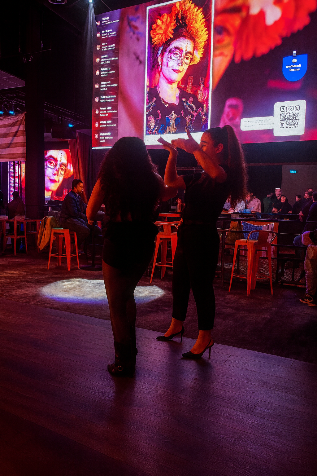 Two people gesturing in a dim event hall before a large screen showing a colorful portrait, comments and a QR code.