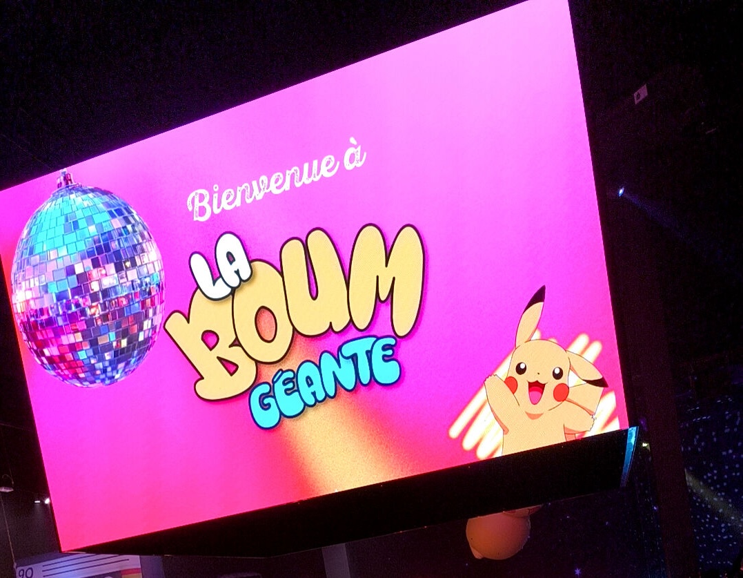 Angled view of a large pink welcome screen reading “Bienvenue à LA BOUM GÉANTE,” with a disco ball and yellow cartoon character in a dark event hall.