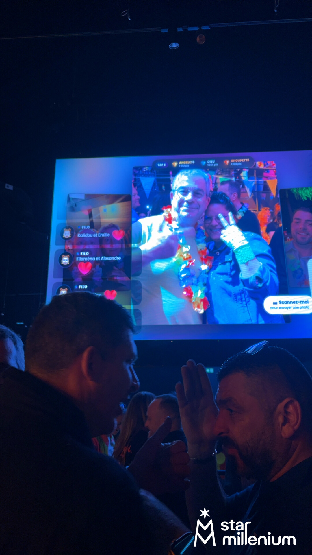 Dos hombres conversan en una sala iluminada en azul ante una pantalla gigante con fotos de participantes, corazones y una invitación para escanear.