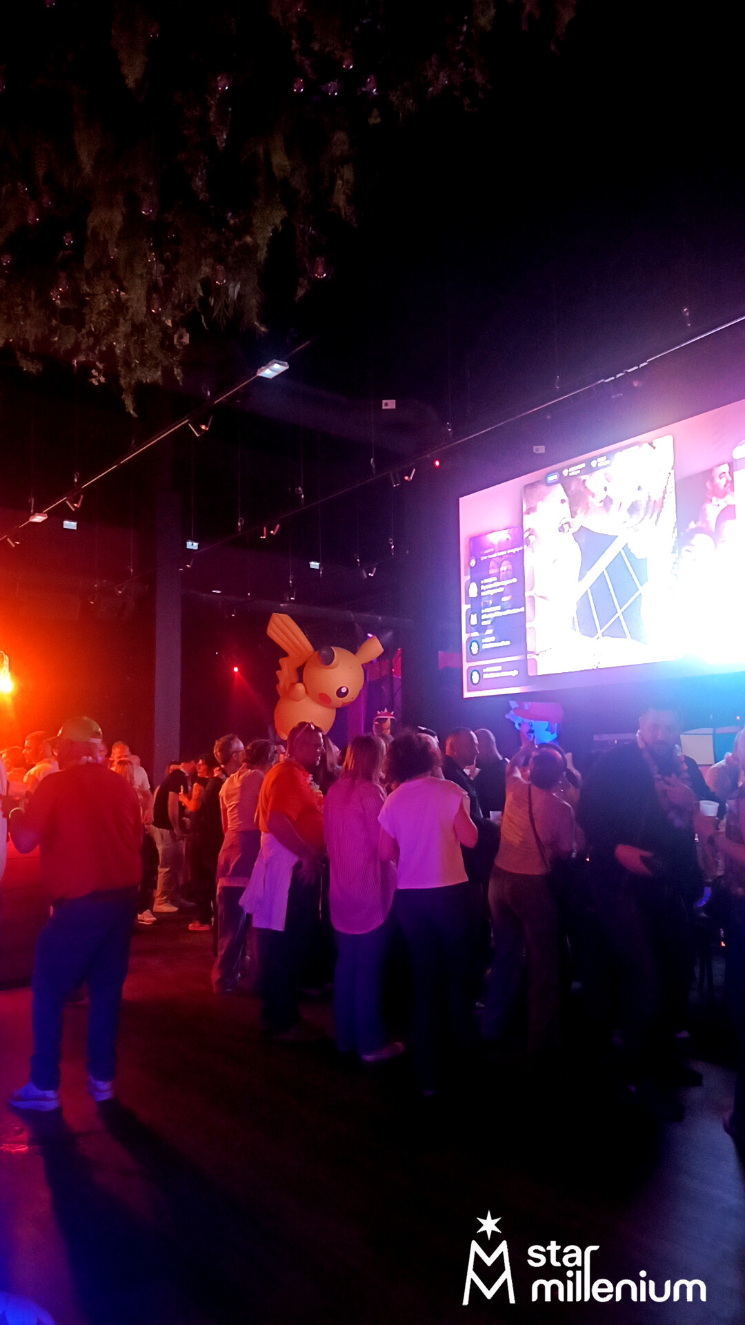 Menschenmenge in einer dunklen Eventhalle mit rotem und violettem Licht, großer Pikachu-Dekoration und hellem Wandschirm