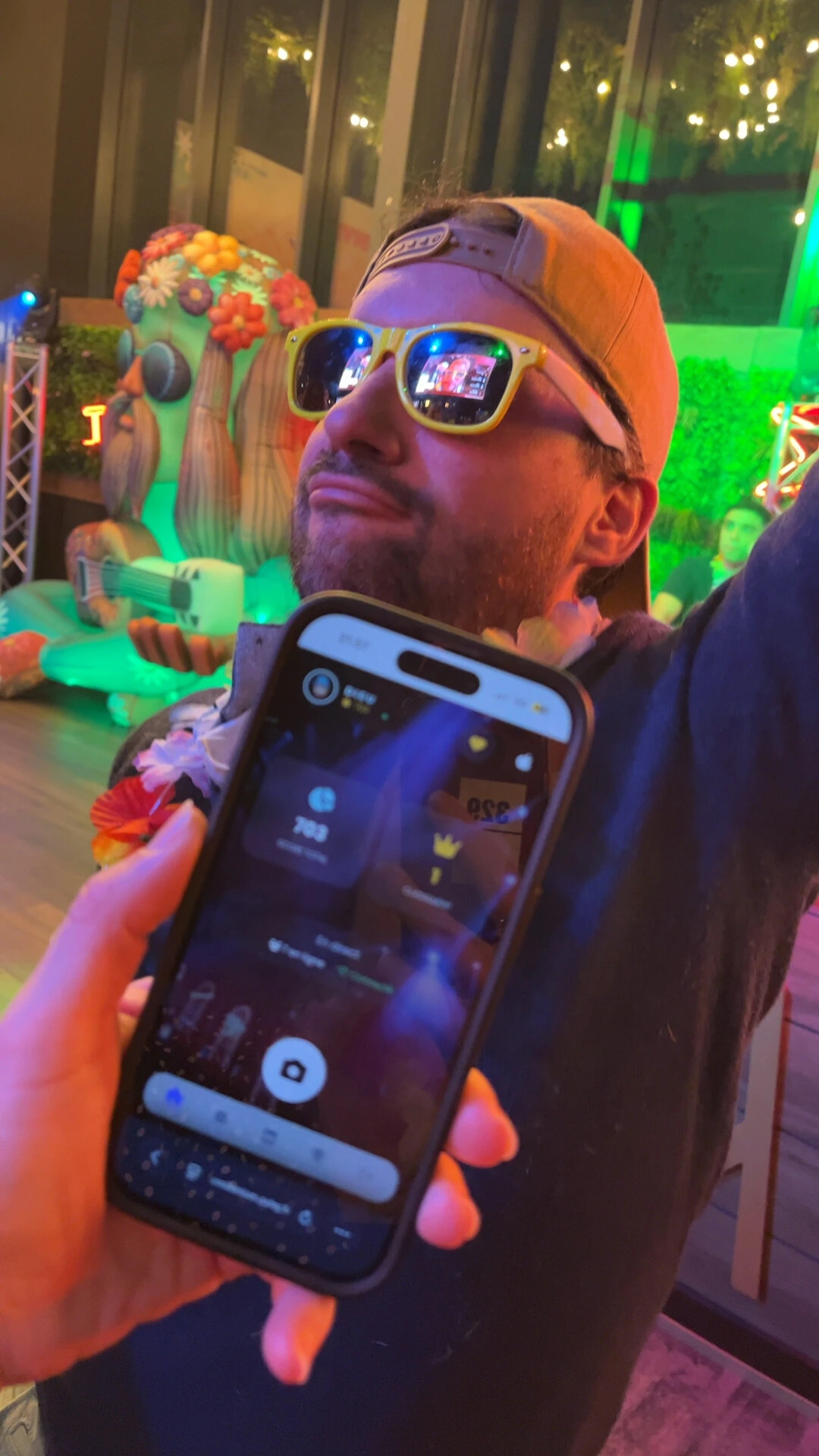 Person in yellow sunglasses and a backwards cap posing in front of a flower-covered cactus while a smartphone is held in the foreground under colorful lights