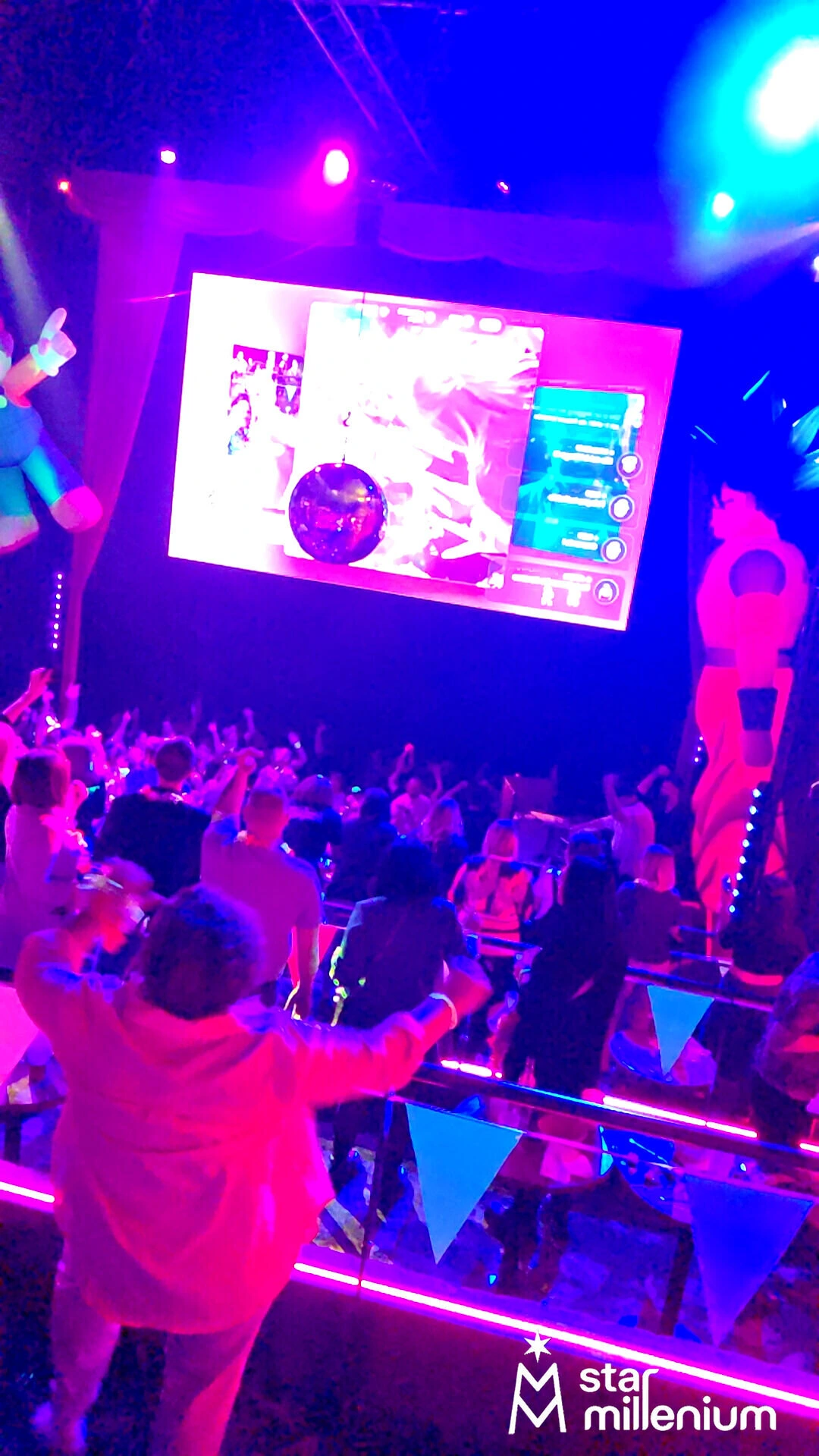 Crowded indoor dance floor under pink, purple and blue neon lights, with guests using phones and a large suspended screen