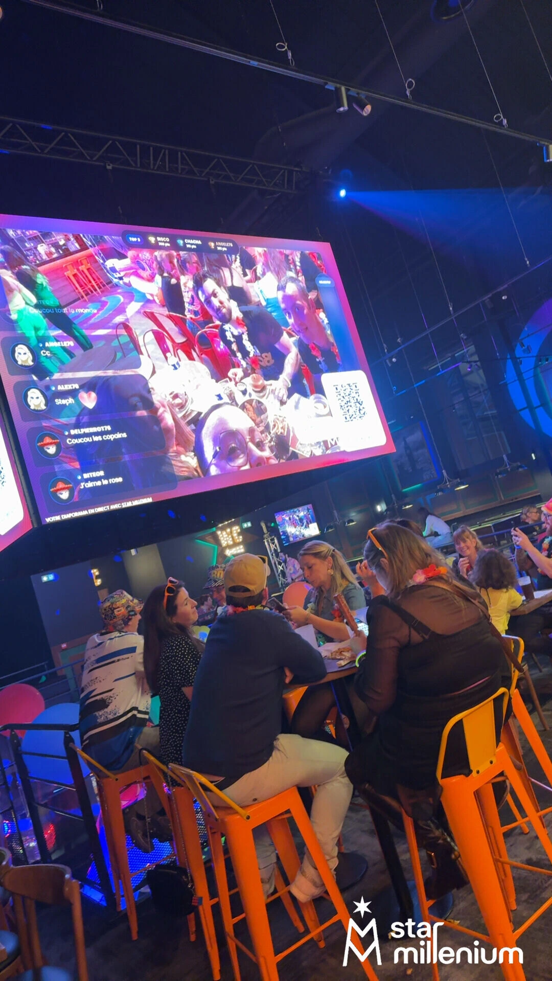 Adults gathered around high tables in a blue-lit indoor venue beneath a giant screen showing shared photos, messages and a QR code.