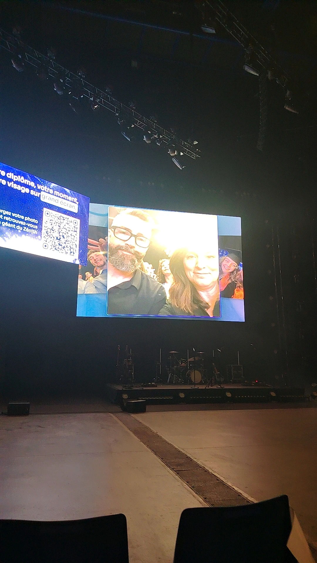Empty indoor stage with a drum kit and lighting rigs, a giant screen showing a group selfie and portrait montage, and a side panel with a QR code