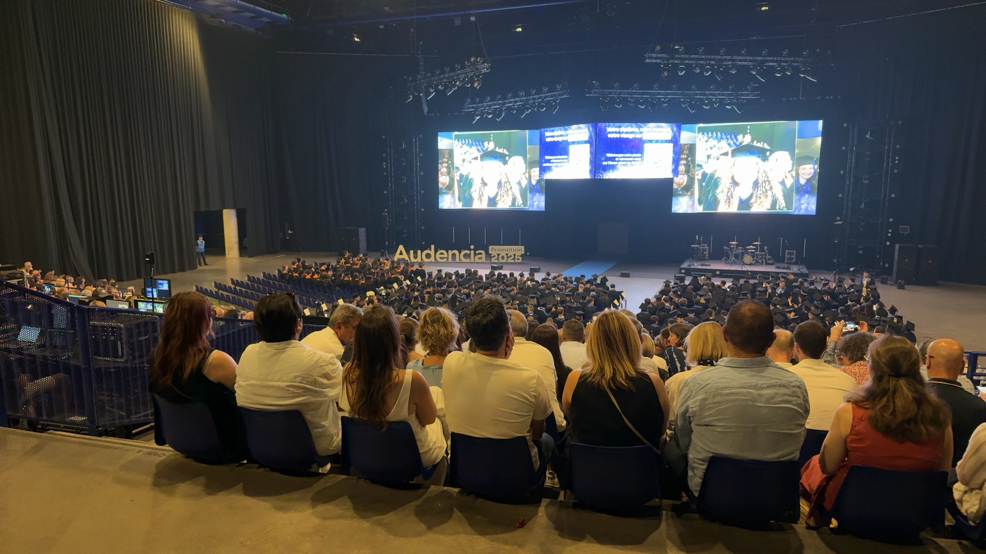 Crowded auditorium with many attendees in graduation caps facing a stage with large screens and musical equipment