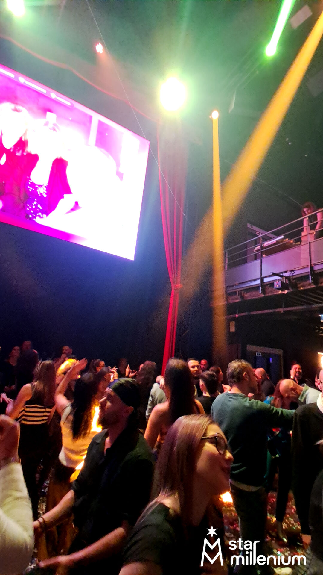 Crowded indoor dance floor under yellow, green and red stage lights, with a bright large screen and technical mezzanine