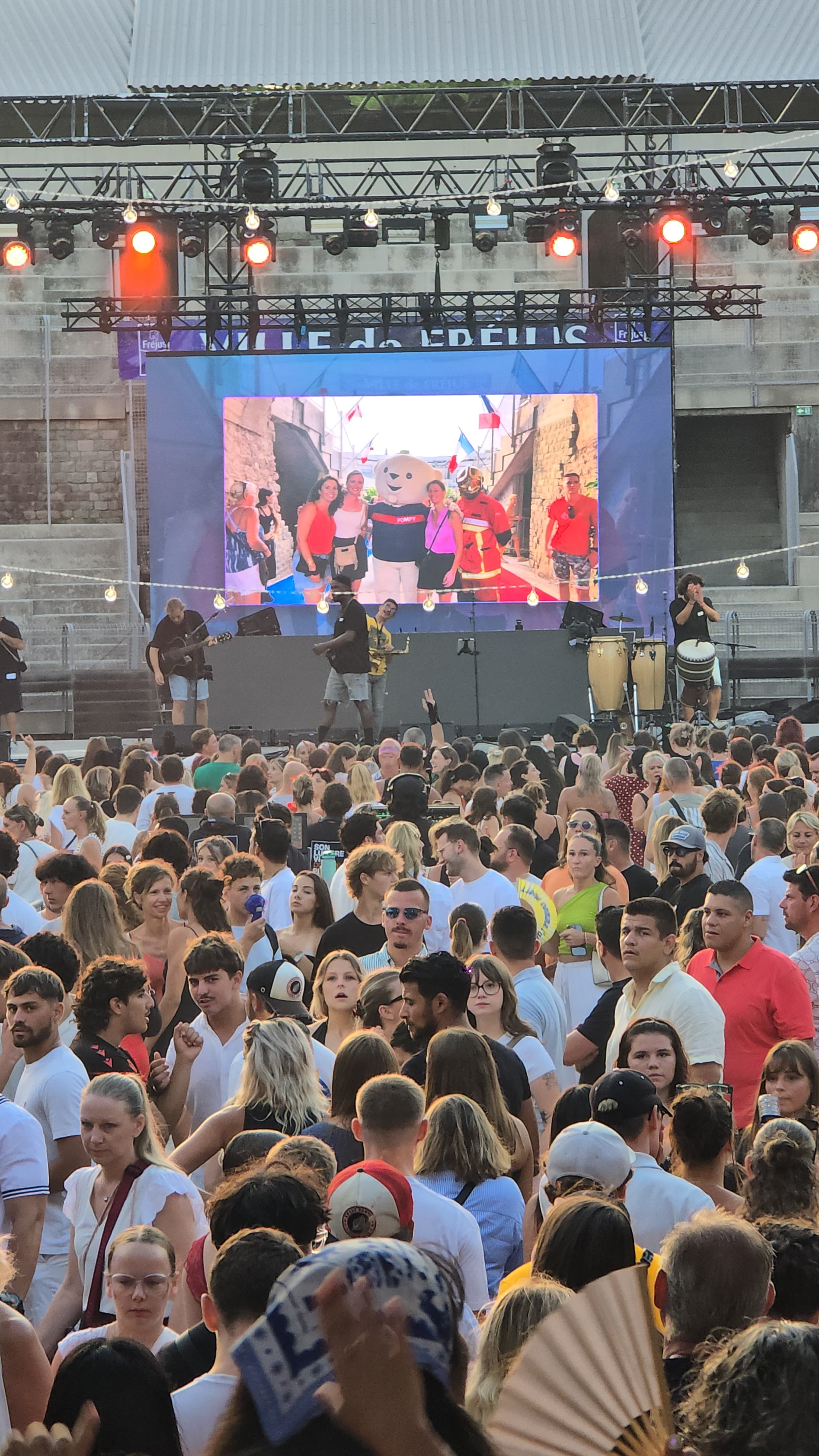 Folla davanti a un palco all’aperto con grande schermo, musicisti e spettatori che usano smartphone