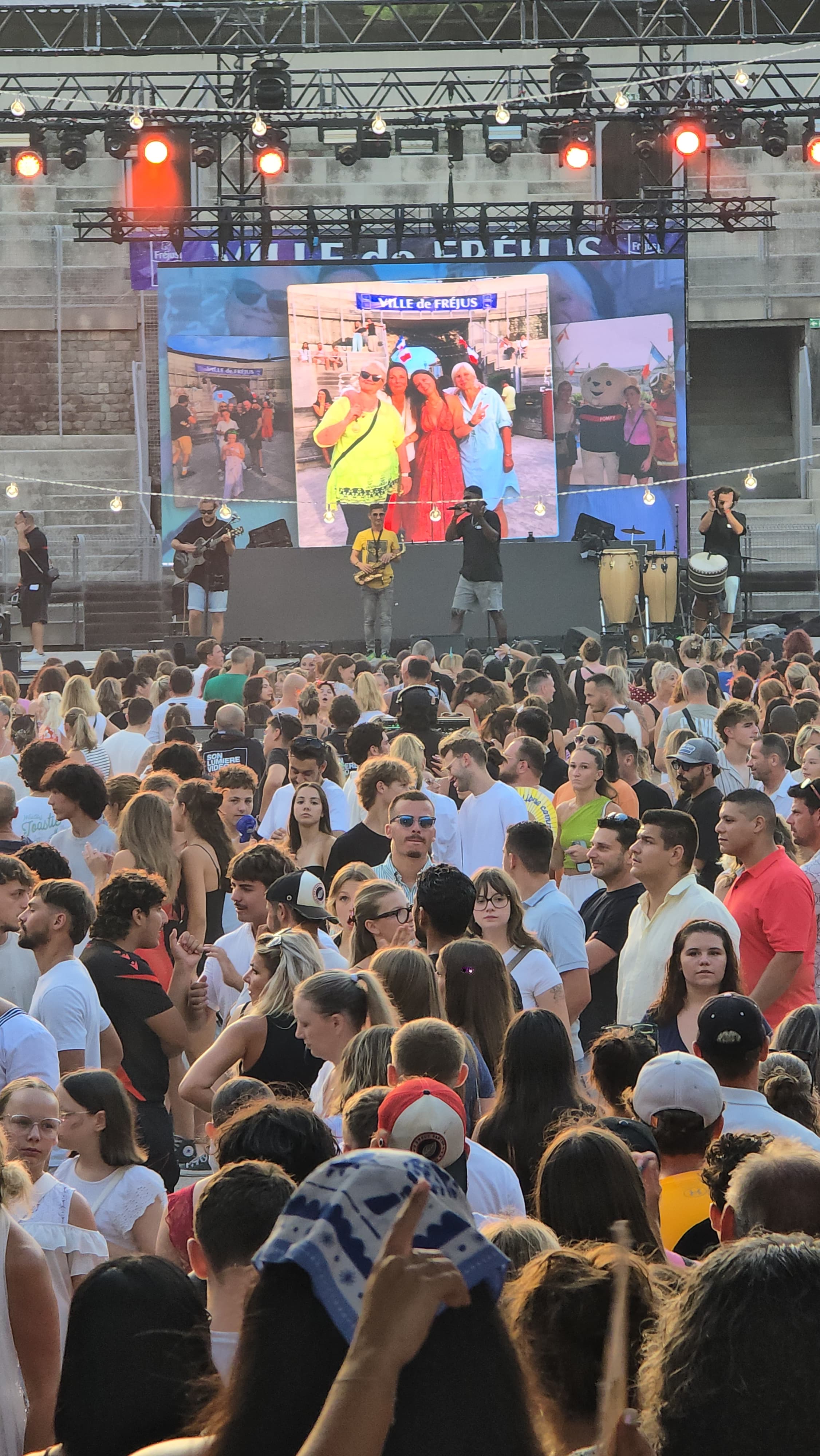 Dichte Menschenmenge vor einer Außenbühne mit Live-Musik, rotem Licht und großem Bildschirm mit mehreren Fotos